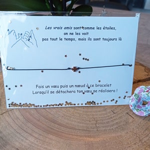 Peut inclure: Un bracelet en fil noir avec un petit charme en forme de trèfle à quatre feuilles. Le bracelet est présenté sur une surface en bois avec une carte contenant du texte français et des confettis en forme d'étoiles.
