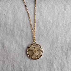 May include: A gold-colored necklace with a delicate floral pendant. The pendant features an openwork design with a central flower shape. The necklace hangs on a white background.