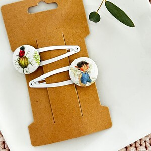 Könnte beinhalten: Zwei weiße Haarklammern mit bunten Designs. Eine Klammer zeigt eine Marienkäfer auf einer Blume und die andere eine Fee mit Flügeln. Die Klammern werden auf einer braunen Karte angezeigt.