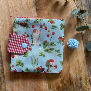 Könnte beinhalten: Eine kleine blaue Stoffbörse mit einer rot-weiß karierten Klappe und einem weißen Knopf. Die Börse hat einen Druck mit Gnomen und Erdbeeren.