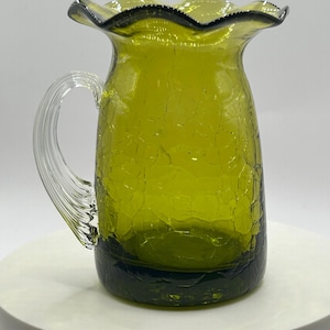 May include: A vibrant olive green glass pitcher featuring a distinct crackle texture throughout the body. The vessel has a clear, ribbed glass handle attached to the side and a wide, ruffled, scalloped rim at the top opening for pouring.