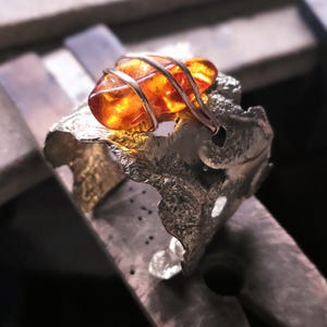 May include: A silver ring with a large amber stone. The amber is a warm orange hue, held in place by silver wire. The ring has a textured, organic design. The ring is likely made in the UK.