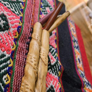 Op de afbeelding: Close-up van drie houten gereedschappen met gesneden details, rustend op een kleurrijk geweven textiel. De gereedschappen hebben lichtbruine handvatten en donkerdere uiteinden. Het textiel heeft een levendig patroon met rode, groene en zwarte tinten.