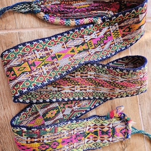 May include: A colorful woven belt with a detailed geometric pattern. The belt features a variety of colors including pink, yellow, green, and blue. The ends of the belt have tassels and a braided cord for tying. The belt is approximately 3 inches wide.