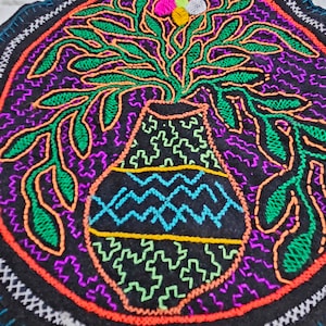 Puede incluir: Un círculo de tela negro con bordado colorido de un jarrón con un arreglo floral. El jarrón está decorado con patrones en zigzag de color azul, verde y amarillo. La flor es rosa, blanca y amarilla.