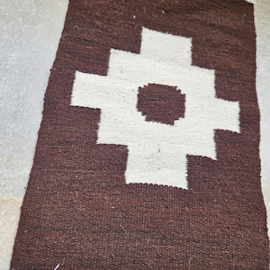 May include: A rectangular, handwoven rug with a brown background and a large white cross-shaped design in the center. The rug has a fringe along one edge and is made of a textured material.