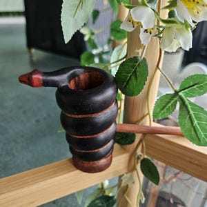 Puede incluir: Una pipa de madera tallada a mano con un diseño de serpiente negra enroscada, cabeza de color marrón rojizo y un tallo largo y delgado. El cuerpo de la serpiente forma el hornillo de la pipa, mostrando vetas de madera natural entre los segmentos oscuros.
