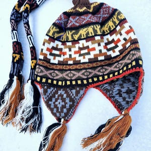 May include: A colorful knitted hat with a long tassel and ear flaps. The hat features a geometric pattern in shades of brown, red, yellow, white, and black.