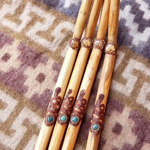 Könnte beinhalten: Vier Holzpfeifen mit aufwendigen Schnitzereien und türkisfarbenen und roten Akzenten. Die Pfeifen sind in einer Reihe auf einem braunen und weißen gemusterten Stoff angeordnet.