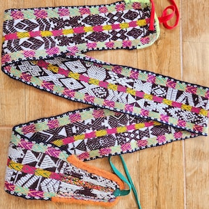 May include: A vibrant woven belt featuring a geometric design in white, brown, pink, yellow, and green. The belt has a black border and red and green ties. The ends of the belt are orange and multi-coloured.