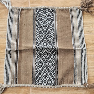 May include: Square woven textile with a brown, black, and white geometric pattern. The central design features a diamond pattern, flanked by vertical stripes. Tassels adorn each corner. The textile is likely a decorative pillow cover or small rug.
