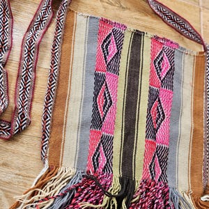 Puede incluir: Un bolso tejido de tapicería colorido con un borde de flecos. El bolso tiene una correa larga con un patrón geométrico. El bolso está hecho de fibras naturales y tiene un aspecto rústico y hecho a mano.