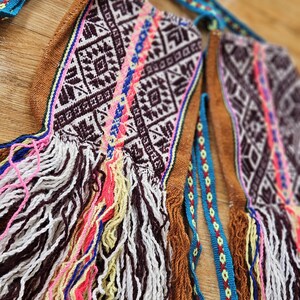 May include: A brown woven bag with colorful fringe and a geometric pattern. The bag has a blue and green strap with a geometric pattern.