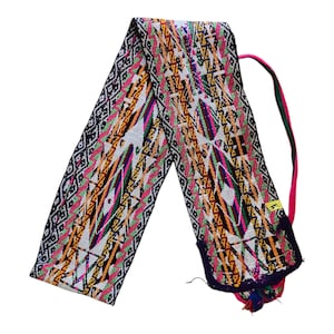 May include: A colorful woven textile with a geometric pattern. The design features black, white, pink, orange, and green threads. The textile has a long, rectangular shape with a pink strap attached.