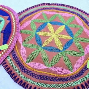 Puede incluir: Dos posavasos redondos de tela bordada con un diseño floral rosa, verde, azul y amarillo. Los posavasos tienen un borde festoneado y están cosidos con una variedad de colores.