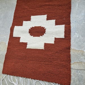 May include: A rectangular, handwoven rug with a rust-colored background and a large white geometric design in the center. The rug has a fringe of white threads along one edge. The rug is likely made of natural fibers.