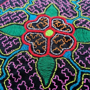 Può includere: Primo piano di un tessuto nero con ricami colorati. Il disegno presenta un grande fiore con punti rossi, arancioni, verdi e gialli. Lo sfondo è pieno di un motivo ripetitivo di punti rosa, blu e gialli.