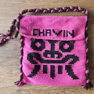 May include: A pink woven shoulder bag with a flap closure. The bag features a black geometric design and the word "CHAVIN" in black letters. The strap and trim are a combination of brown and pink yarn.