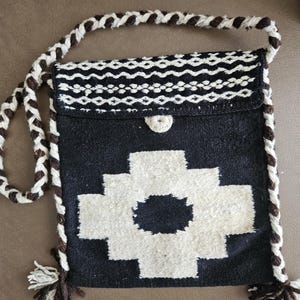 May include: A black and white woven crossbody bag with a geometric design. The bag features a white cross-like pattern on the front, a flap closure, and a braided strap with tassels. The bag is approximately 10 inches wide and 10 inches tall.