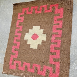 May include: A small, square woven rug with a brown background. A pink geometric border surrounds a central design of a white cross-like shape with a pink center. The rug has a textured appearance and a handmade aesthetic.