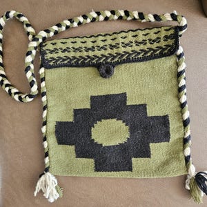 May include: A woven shoulder bag in olive green with a black geometric design. The bag features a braided strap in black, white, and olive green, with tassels at the bottom. The flap is secured with a button.