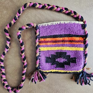 May include: A small, woven shoulder bag with a braided strap. The bag features a purple background with horizontal stripes of orange, yellow, and black. A black geometric design is centered on the front. The strap is a mix of black, tan, and pink.