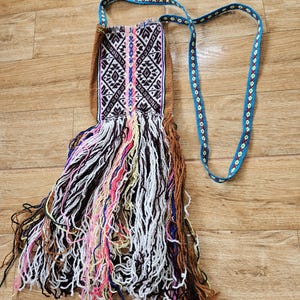 Könnte beinhalten: Eine kleine, braune, gewebte Tasche mit einem langen, bunten Fransen. Die Tasche hat ein geometrisches Muster und einen blauen und gelben Riemen.