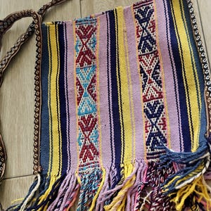 May include: A colorful woven shoulder bag with a striped pattern in shades of blue, yellow, and purple. The bag features intricate geometric designs and a fringe detail at the bottom. The strap is a patterned brown.