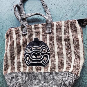 May include: A tote bag featuring a woven pattern of vertical brown and cream stripes with a black herringbone base. The front displays a black embroidered patch of a stylized creature face with prominent teeth and circular eyes, and two handles.
