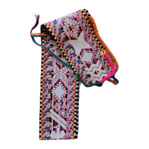May include: A colorful, woven textile belt with intricate geometric patterns in white, purple, orange, pink, and green. The belt has a rectangular shape with a decorative edge and tassels. The textile is likely handmade.
