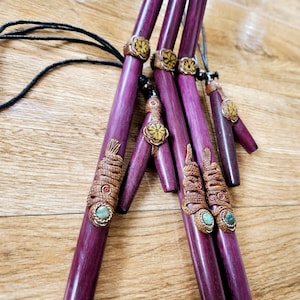 Op de afbeelding: Vijf paarse houten pijpen met ingewikkelde houtsnijwerk en kleurrijke edelsteenaccenten. De pijpen zijn aan elkaar geregen met een zwart koord en hebben decoratieve bloem- en slangontwerpen.