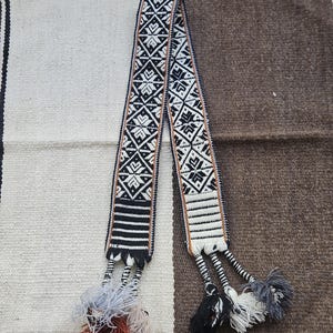 May include: A woven belt with a geometric pattern in black, white, and brown. The belt has tassels at both ends, with a mix of black, white, gray, and brown threads. The background is a woven textile in shades of white and brown.