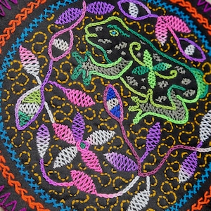 May include: A circular embroidered patch with a black background and colorful stitching. The design features a stylized frog with green, gray, and white stitching, surrounded by pink, purple, and white floral motifs. The outer edge of the patch is a multi-colored border.