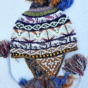 Könnte beinhalten: Eine bunte Strickmütze mit geometrischem Muster und weißen Perlen. Die Mütze hat einen braunen Bommel oben und lange Quasten an den Seiten.