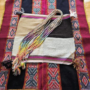 May include: A woven textile with a geometric pattern in red, black, and teal. A smaller, multi-colored woven bag with a rainbow-colored strap is on top. The bag has a tan, black, and gray color scheme. Tassels are on the corners of the textile.