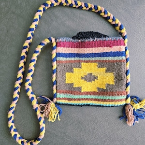 May include: A small, woven shoulder bag with a colorful geometric pattern. The bag features a yellow cross design on a gray background, with stripes of pink, blue, and brown. The strap is braided in blue, yellow, and pink.