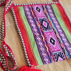 May include: A colourful woven bag with a red strap and a red tassel. The bag has a geometric pattern with stripes of red, green, pink, blue and yellow.