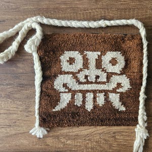 May include: A brown woven bag with a white geometric design and a white rope strap. The bag is rectangular with a textured surface. The strap is thick and braided.