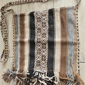 May include: Handwoven shoulder bag with a striped pattern in shades of brown, black, and beige. The central panel features a repeating geometric design in brown and white. The bag has a fringed bottom and a woven strap.