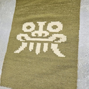 May include: A rectangular olive green woven rug with a cream-colored geometric design in the center. The design appears to be a stylized face or figure. The rug has a textured surface and a simple, minimalist aesthetic.