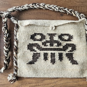 May include: A woven shoulder bag with a geometric design in brown on a cream background. The bag has a braided strap in brown and cream. The bag is rectangular with a flap closure and a button.