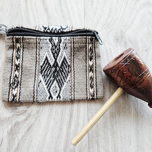 May include: A small, rectangular, woven pouch with a black zip and a patterned design in shades of grey, black, and white. Beside it is a carved wooden pipe with a light-coloured stem and intricate details.