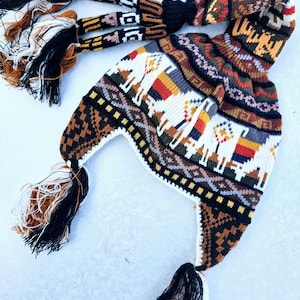 Puede incluir: Un gorro de punto blanco con un patrón geométrico colorido con llamas. El gorro tiene una borla larga negra y dos borlas más cortas a los lados. El gorro está hecho de lana y es perfecto para el clima frío.