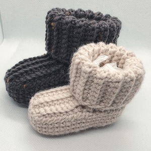 May include: Two handmade baby booties. One is dark grey with flecks of colour, the other is a light beige. Both are made of a thick, textured yarn and have a folded-over cuff. The booties are designed to keep a baby's feet warm.