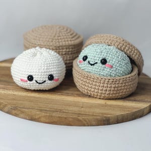 May include: Two crocheted plush toys shaped like steamed buns. One is white with black eyes and pink cheeks. The other is light green with black eyes and pink cheeks, nestled inside a tan crocheted container with a matching tan lid nearby.
