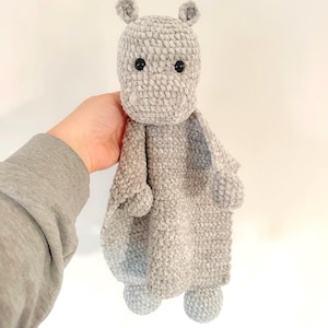May include: A gray crocheted hippopotamus baby blanket with a soft, plush texture. The blanket has a small hippopotamus head with black eyes and a gray body with pom-poms on the feet.
