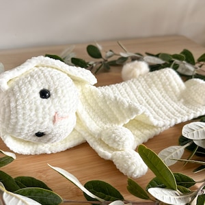 May include: A white crocheted bunny rabbit toy with a long body and a small tail. The bunny is lying on a wooden surface with green leaves around it.