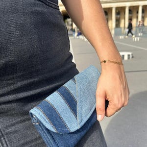 Peut inclure: Une pochette en jean avec fermeture à rabat. La pochette présente un motif patchwork avec différentes nuances de denim bleu et des coutures jaunes contrastantes. La personne porte un bracelet en or et une combinaison en jean noir.