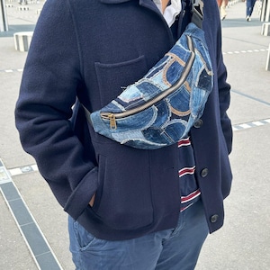 Op de afbeelding: Een denim heuptas met een patchwork ontwerp. De tas heeft een gouden ritssluiting. De tas wordt over het lichaam gedragen. De persoon draagt een marineblauwe jas en een blauwe broek.