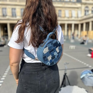 Peut inclure: Un sac à dos rond en jean avec un motif patchwork dans différentes nuances de bleu. Le sac à dos a une poignée supérieure et des bretelles réglables. Le sac est porté sur le dos. L'arrière-plan est une scène extérieure floue.
